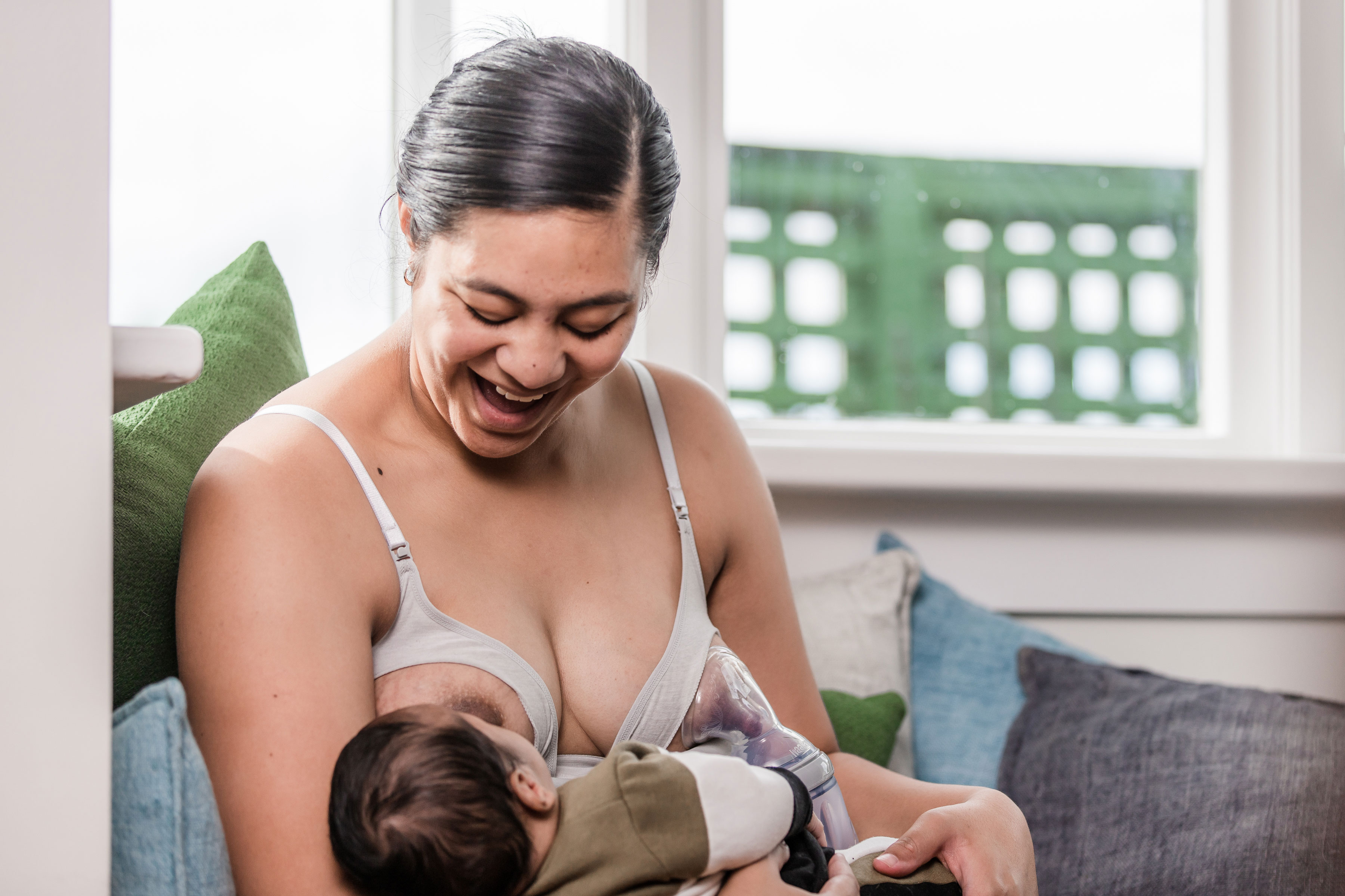 Fröhliche Mutter stillt ihr Baby an der rechten Brust und pumpt an der linken Brust Milch mit einer Haakaa Milchpumpe (Gen 3) ab.